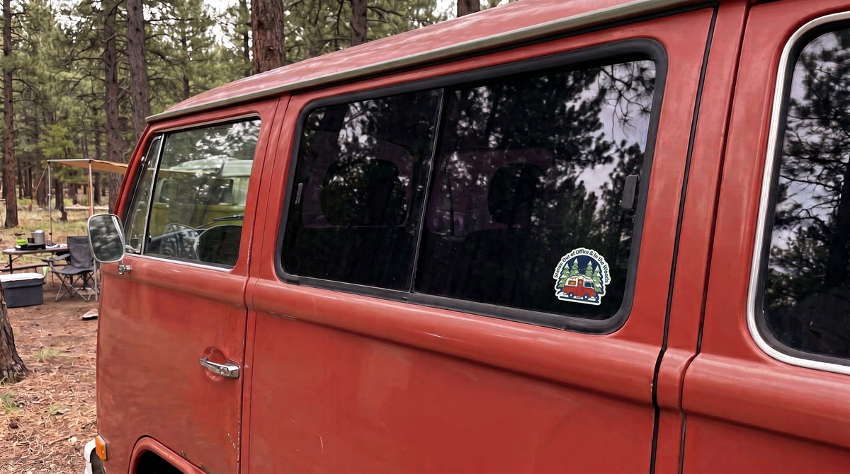 Out of Office & In the Woods sticker from the camping bundle on a red camper van window at a forest campsite