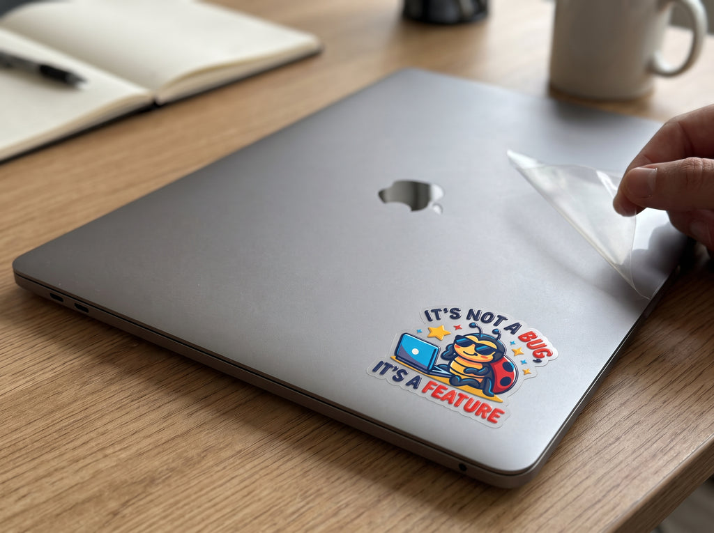 It's Not a Bug, It's a Feature waterproof sticker on a gray laptop lid next to a notebook and coffee mug