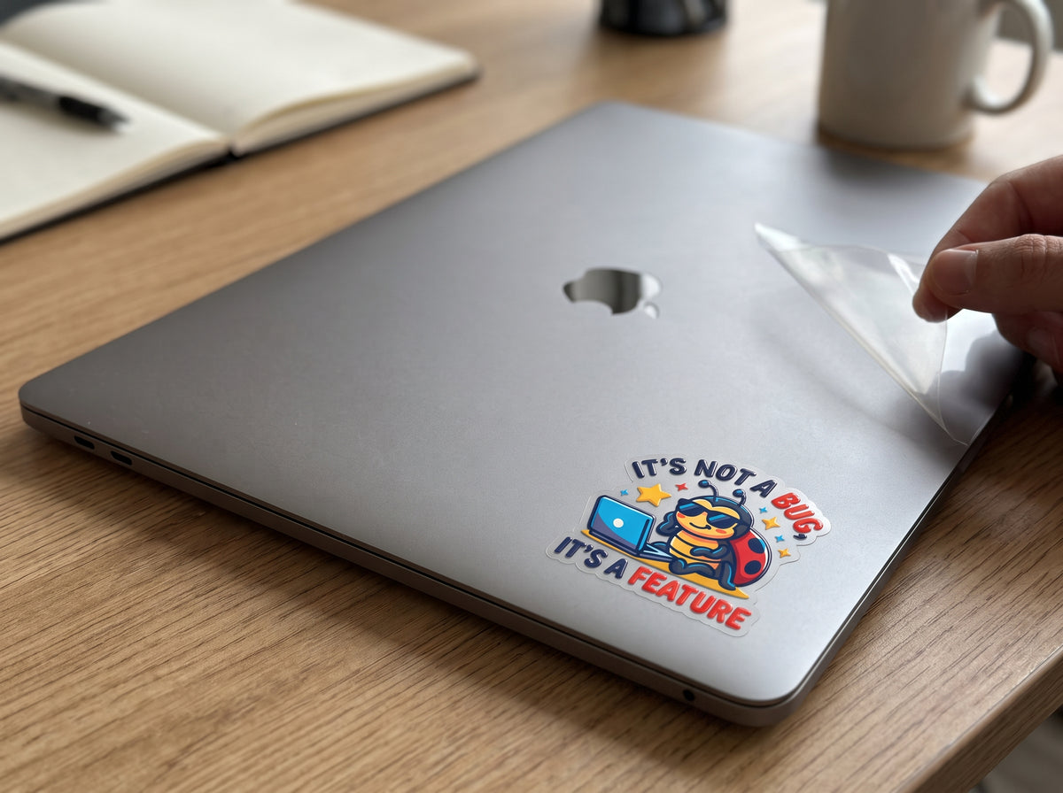 It's Not a Bug, It's a Feature waterproof sticker on a gray laptop lid next to a notebook and coffee mug