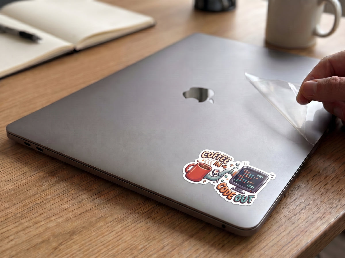 Coffee In, Code Out waterproof sticker on a gray laptop lid next to a notebook and coffee mug